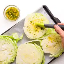 A hand brushes dressing over cabbage steaks on a prepared pan.