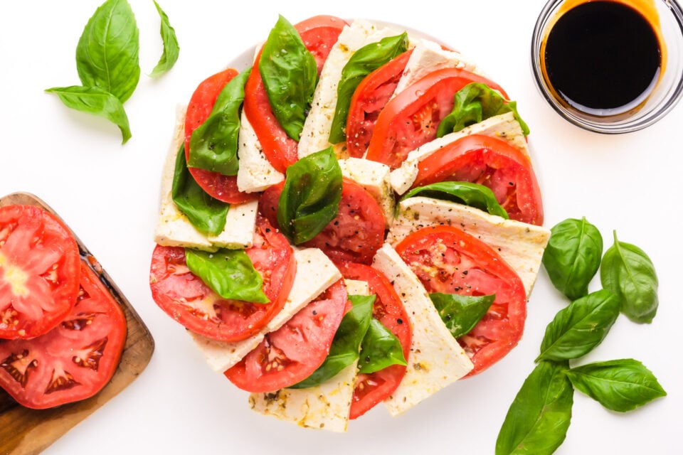 Looking down on a layered caprese salad with fresh tomatoes, basil leaves, and tofu. There are cut tomatoes, basil, and a bowl of balsamic glaze nearby.