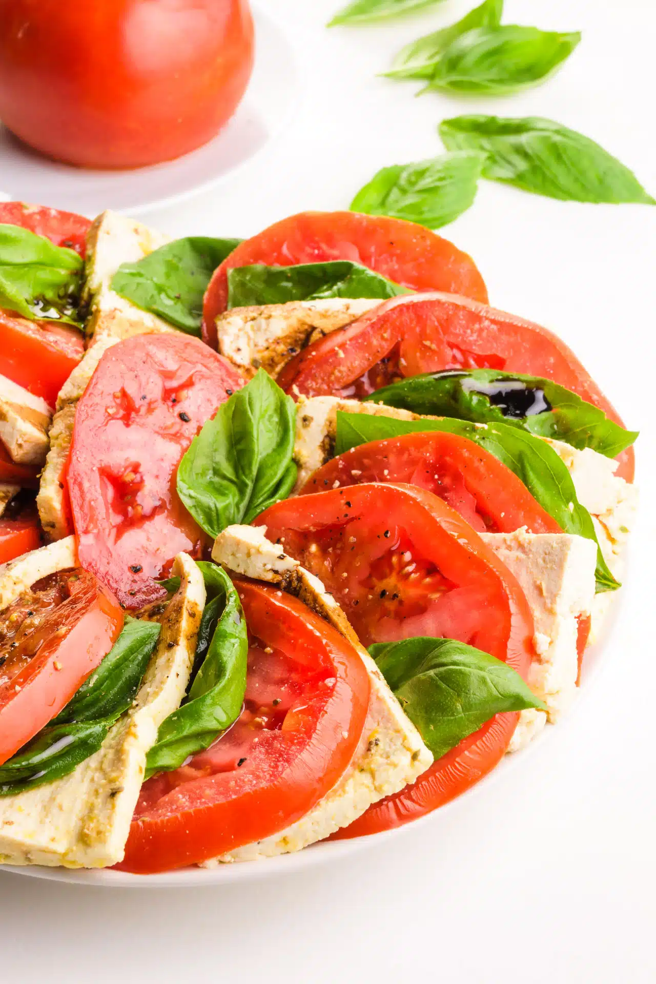 A closeup of vegan cpreses salad on a plate. There are fresh basil leaves and a tomato in the background.