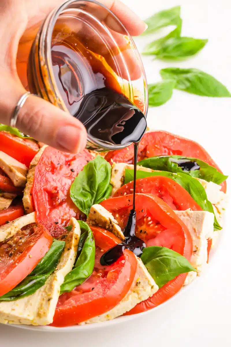 A hand holds a small bowl of balsamic glaze, pouring it over a Caprese salad.