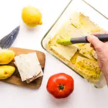 A hand holds a pastry brush, brushing marinade on tofu slices in a small dish. There are lemons and tomatoes and a cutting board nearby.