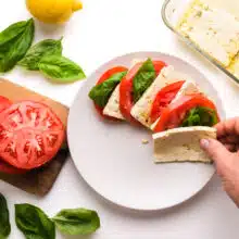A hand places tofu on a plate with sliced tomatoes and basil leaves. There is more tofu in a dish, fresh basil leaves, a lemon, and sliced tomatoes nearby.