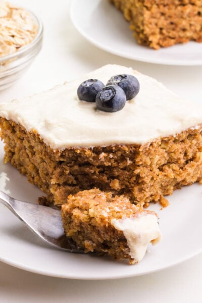 A slice of cake on a plate has a forkful of cake in front of it. There is a bowl of oats and another slice in the background.