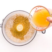 A hand holds a measuring cup, pouring liquid ingredients from a pyrex measuring cup into a food processor with flour ingredients.