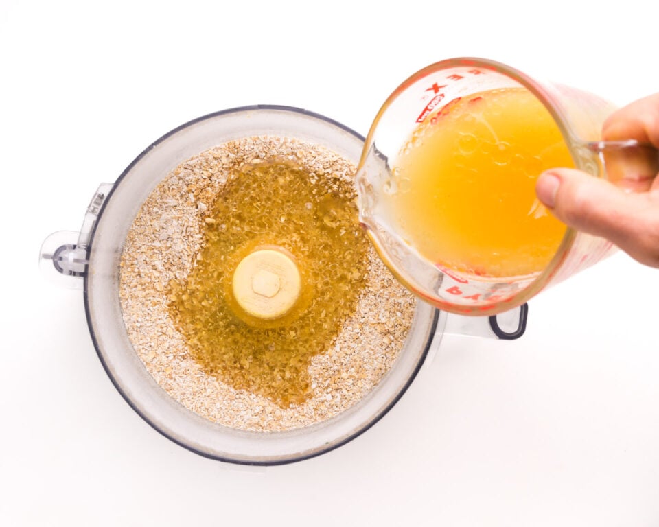 A hand holds a measuring cup, pouring liquid ingredients from a pyrex measuring cup into a food processor with flour ingredients.