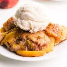 A slice of vegan peach cobbler sits on a plate. It has vanilla ice cream on top.