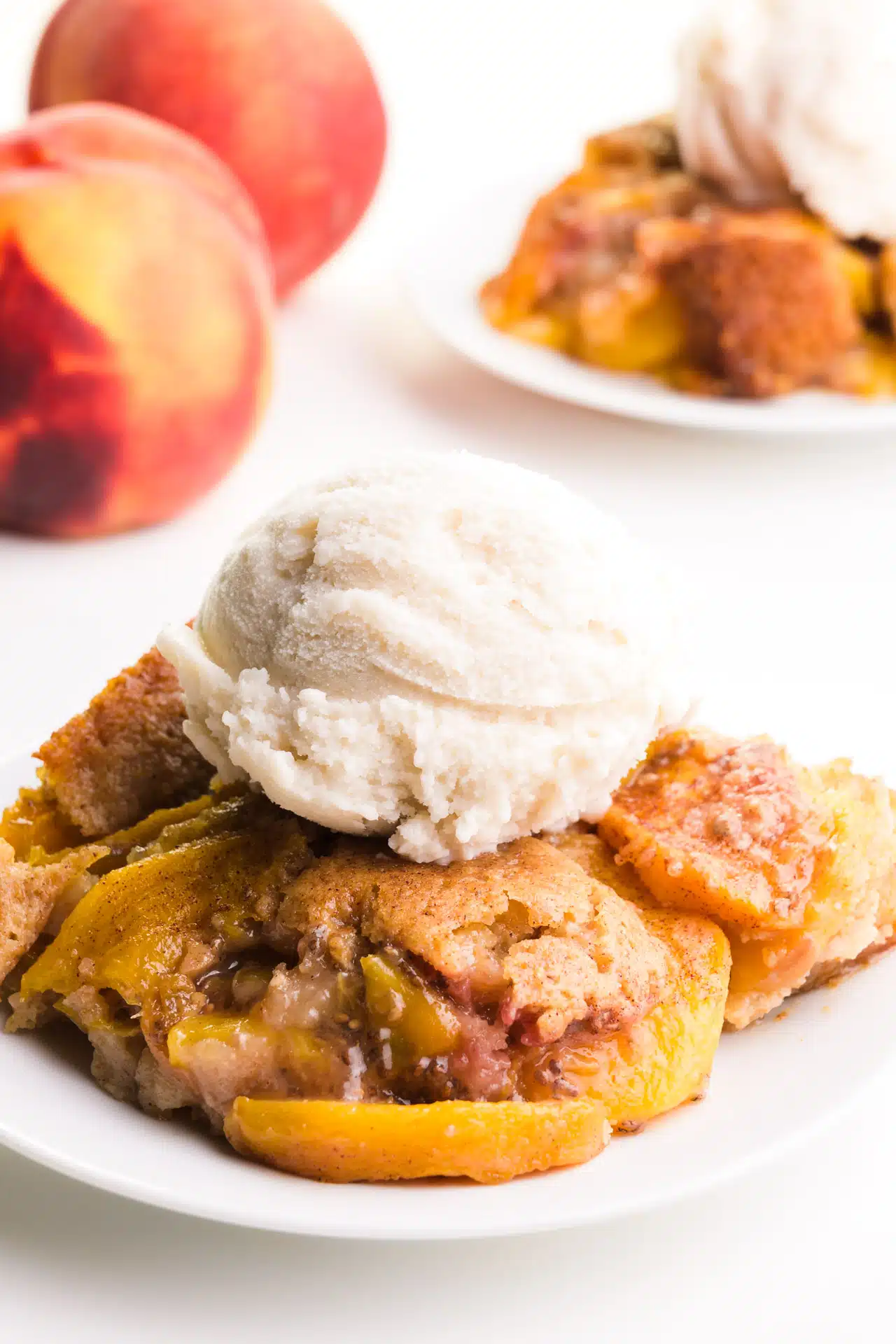 Vegan peach cobbler on a plate sits in front of fresh peaches and another plateful of cobbler.