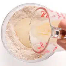 A hand holds a pyrex measuring cup, pouring liquid into a bowl with flour.