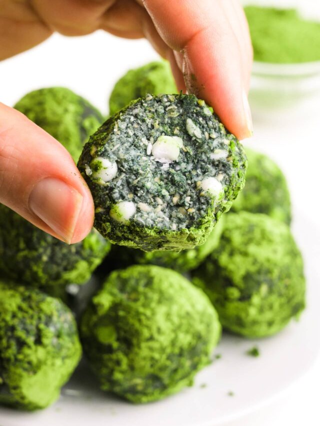 A hand holds an energy ball with a bite taken out, revealing white chocolate chips inside. There is a plate with more energy balls and a bowl of matcha powder in the background.
