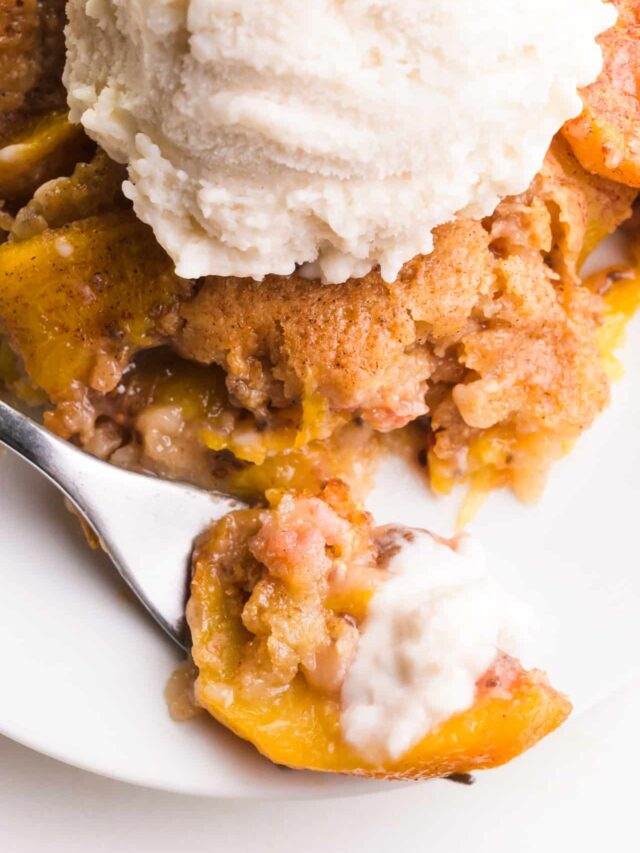 A forkful of vegan peach cobbler sits in front of the rest of the cobbler on a plate. It's topped with vegan vanilla ice cream.