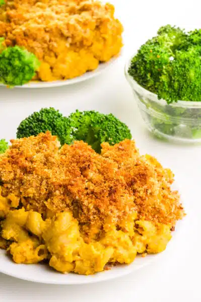 Chickpea Mac and Cheese sits on a plate next broccoli. There is another plate in the background and a bowl of steamed broccoli.