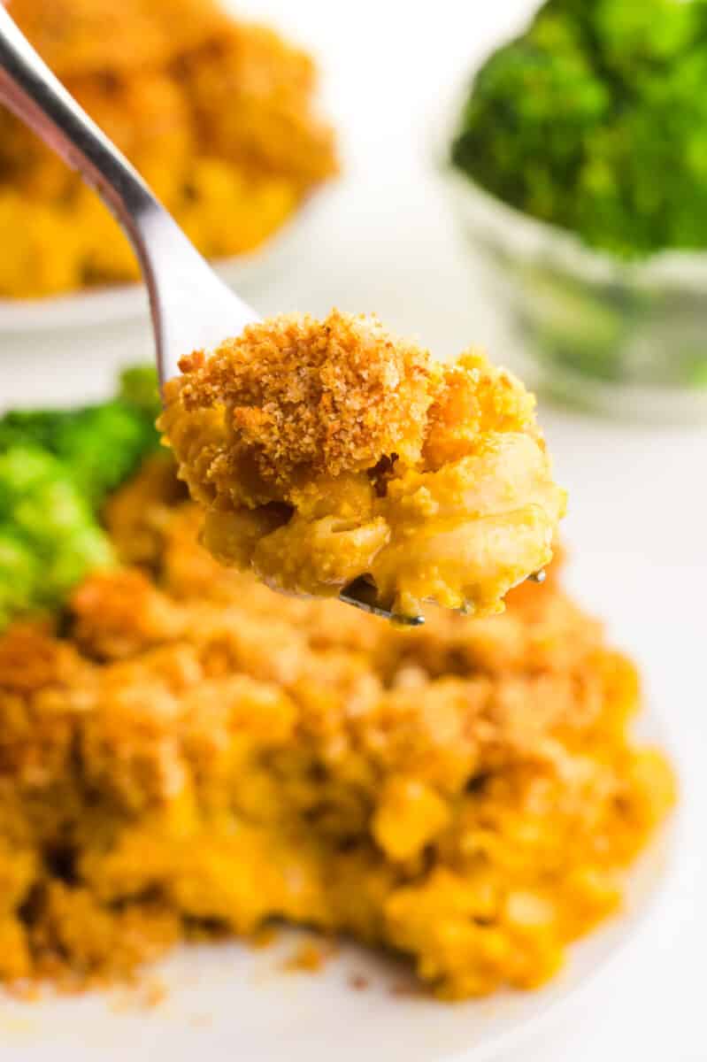 A spoonful of chickpea macaroni and cheese hovers over a plateful. There is more on a plate in the background and a bowl of steamed broccoli.