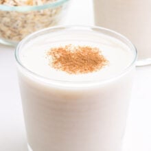 A glass of oat milk sits in front of another glass and a bowl of oats.