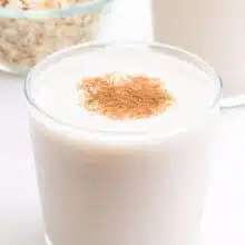 A glass of oat milk sits in front of another glass and a bowl of oats.