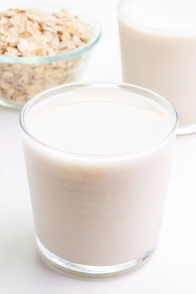 A glass of oat-based milk sits in front of another glass and a bowl of oats.