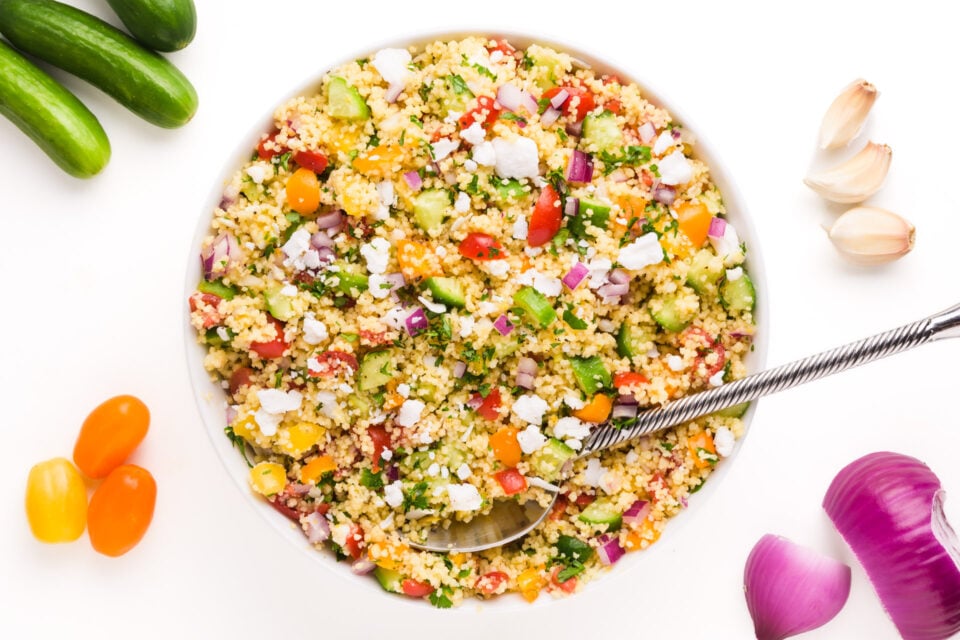 Looking down on couscous salad surrounded by ingredients liked cucumbers, cherry tomatoes, red onions and garlic cloves.