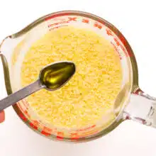 A hand holds a spoonful of olive oil over couscous in a measuring cup.