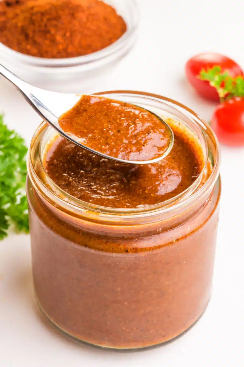 A spoonful of vegan enchilada sauce hovers over a jar full of it. There are tomatoes, herbs, and seasonings behind it.