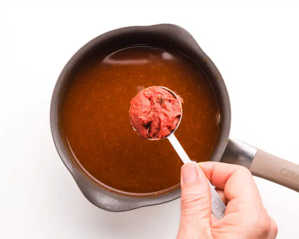 A hand holds a measuring spoon full of tomato paste over a sauce pan with enchilada sauce.