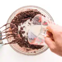 Milk is being poured into a bowl with chocolate cookie batter. There is a mixer sitting by the bowl.