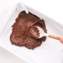 A spatula spreads chocolate batter into a white baking pan.