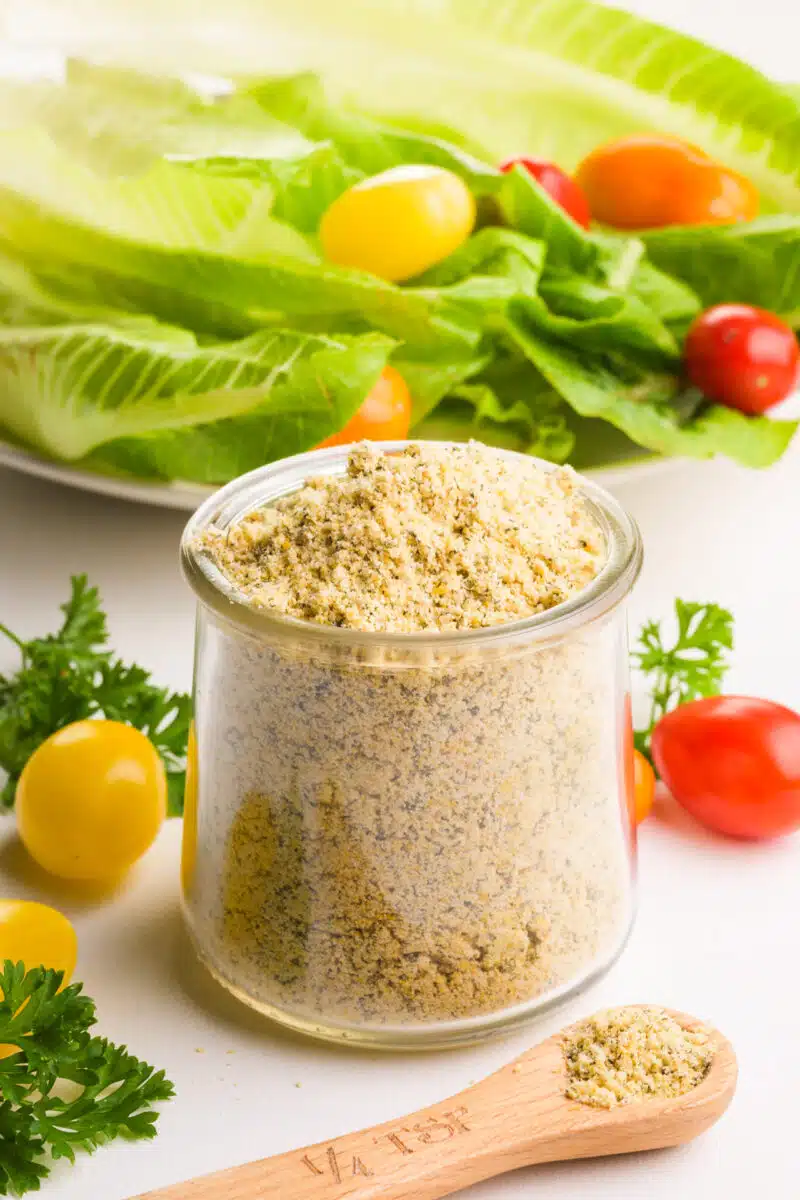 A bowl of vegan ranch seasoning sits on a table with fresh veggies and salad ingredients around it.