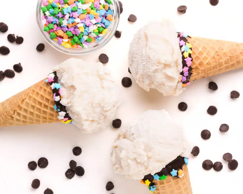 Looking down on three ice cream cones with vanilla ice cream scoops. There are chocolate chips and colorful sprinkles around the cones.