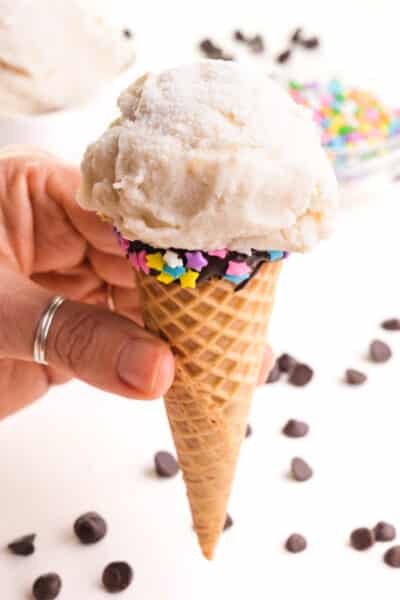 A hand holds an ice cream cone with a scoop of vegan vanilla ice cream.