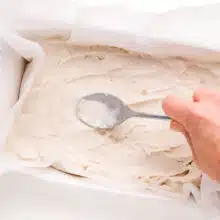 Vanilla ice cream is being spread in a pan lined with parchment paper.