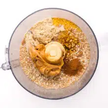 Looking down on a food processor with ingredients, such as oats, cinnamon, syrup sweetener, and peanut butter.