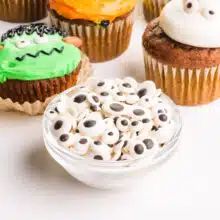 A bowl of vegan googly eyes sits in front of decorated cupcakes.