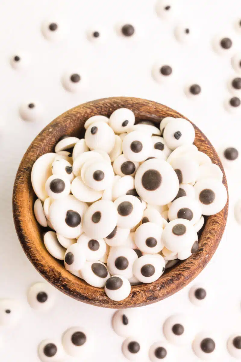 A small wooden bowl is full of edible candy eyes. There are more of the candy eyes scattered around the bowl.