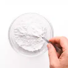 A hand holds a spoonful of cornstarch, getting read to pour it into a bowl with powdered sugar.