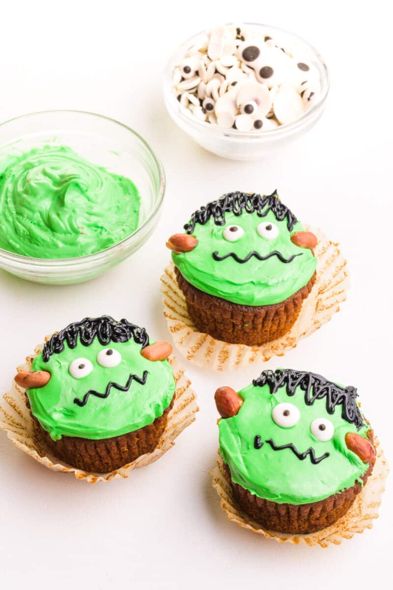 Three Frankenstein cupcakes it next to a bowl of green frosting and googly candy eyes.
