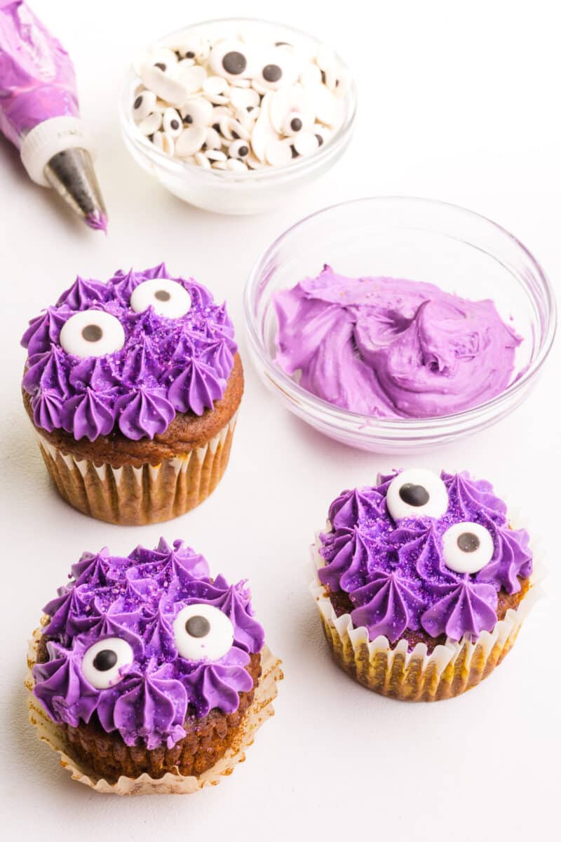 Purple monster cupcakes have googly eyes. They sit next to a bowl of purple frosting and other toppings.