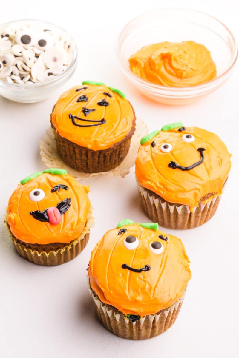Jack-o-lantern cupcakes sit next to a bowl of orange frosting and vegan candy eyes.