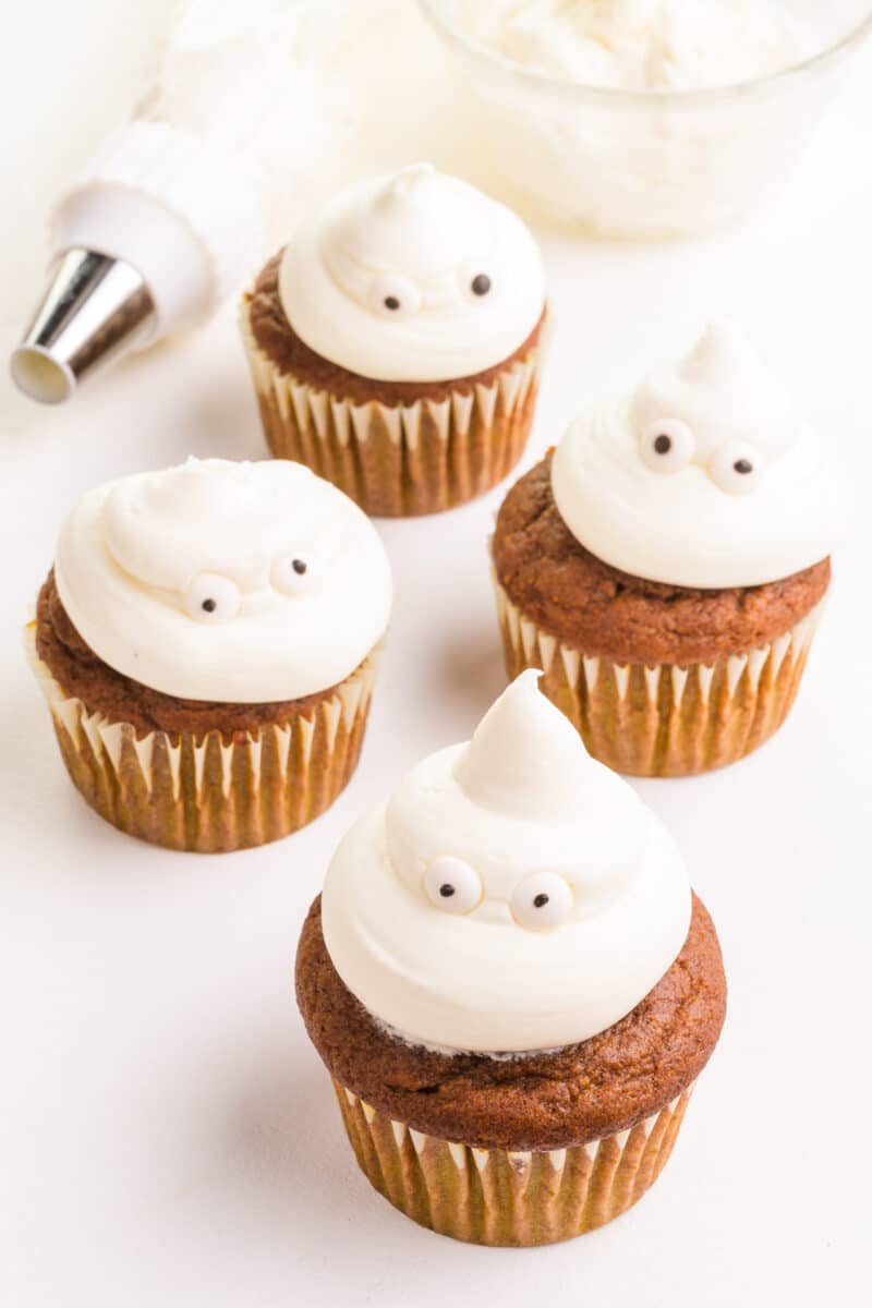 Four ghost cupcakes sit next to a piping bag with white frosting and a bowl of candy eyes.