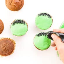 A hand pipes black frosting on green cupcakes. Some plain cupcakes sit beside them.