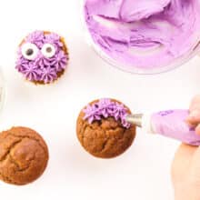 A hand pipes purple frosting stars on cupcakes. One has candy eyes on it and it sits next to a bowl of purple frosting.