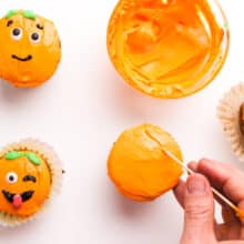 A hand uses a toothpick to etch lines in orange frosting on a cupcake to make it look like a pumpkin.