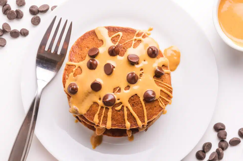 Looking down on protein pancakes with peanut butter and chocolate chips on top. There is a fork beside the pancakes on the plate.