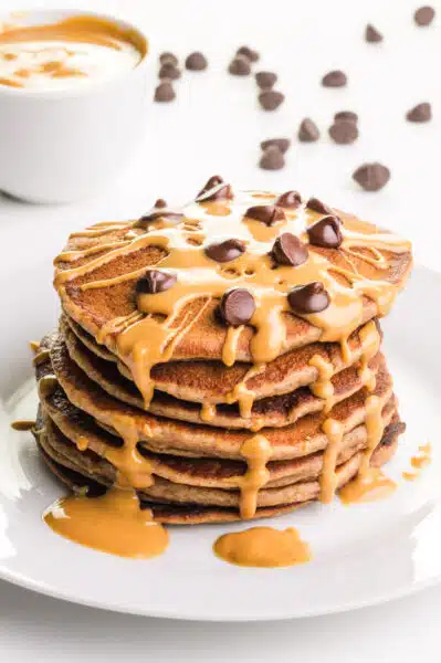 A stack of pancakes has peanut butter and chocolate chips on top. There is a bowl of peanut butter and more chocolate chips in the background.