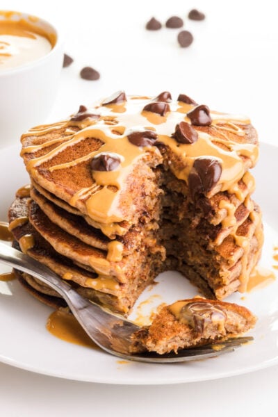 A fork holds a bite of pancake sitting in front of a stack of pancakes with a bite taken out.