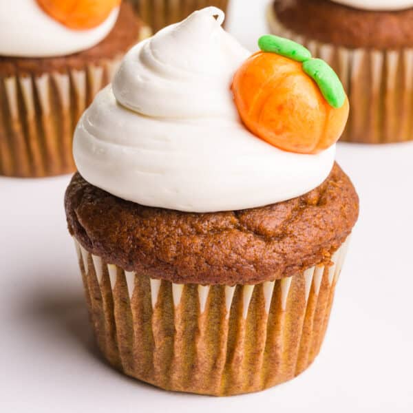 A vegan pumpkin cupcake has white frosting on top and pumpkin candy on the side.