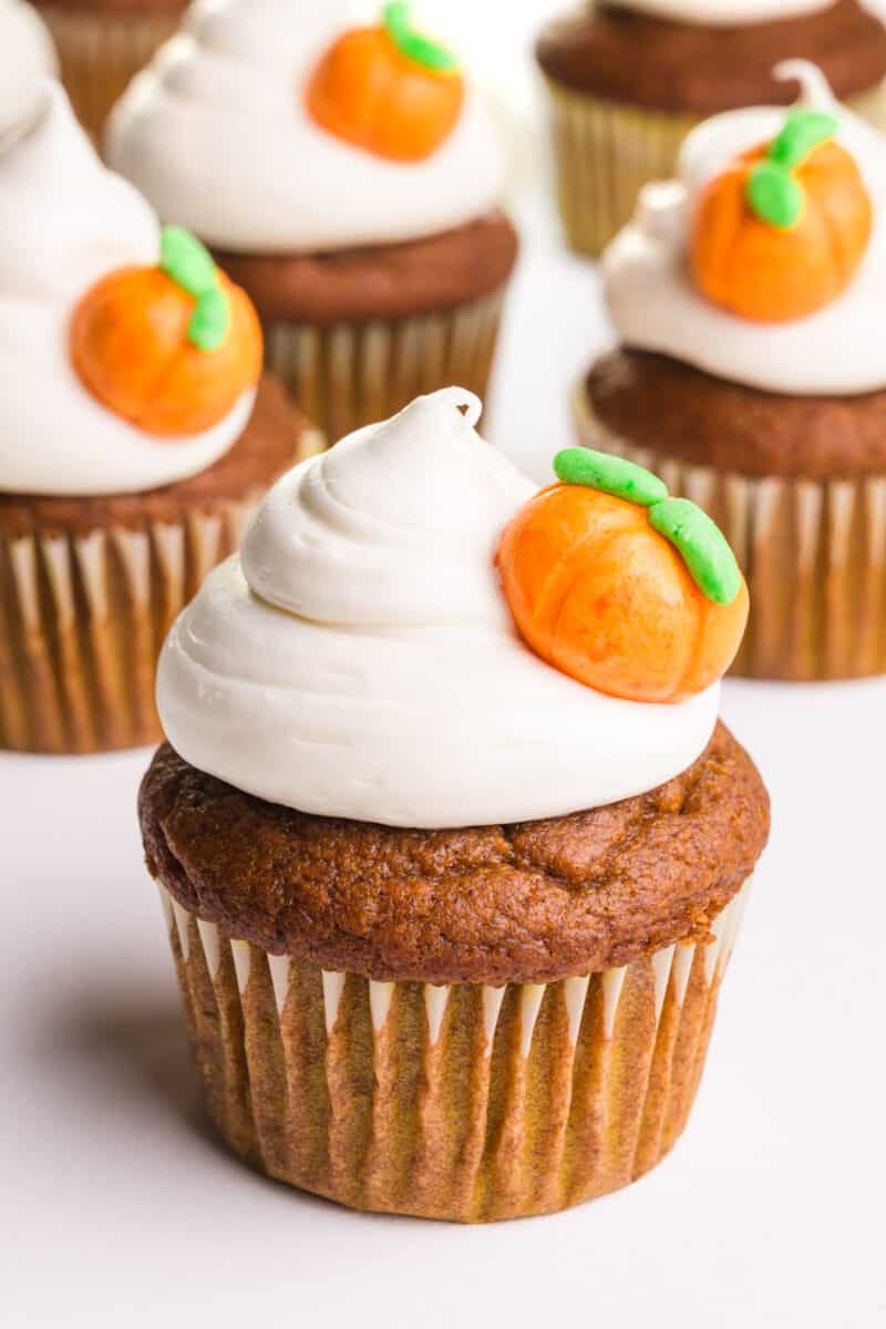 A bunch of vegan pumpkin cupcakes have white frosting on top and pumpkin candies on the side.