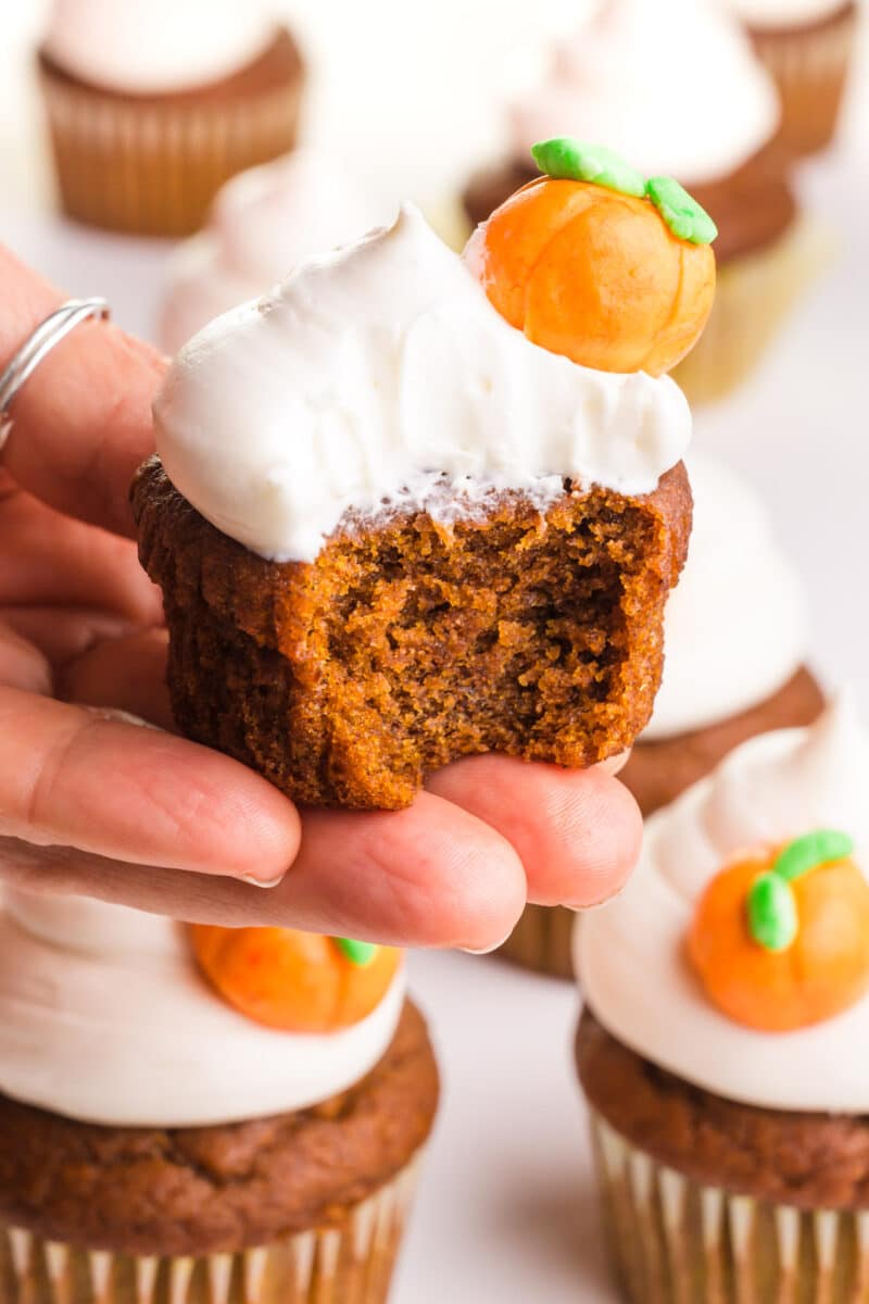 A hand holds a pumpkin cupcake with a bite taken out. It has white frosting on top and a candy pumpkin on the side.