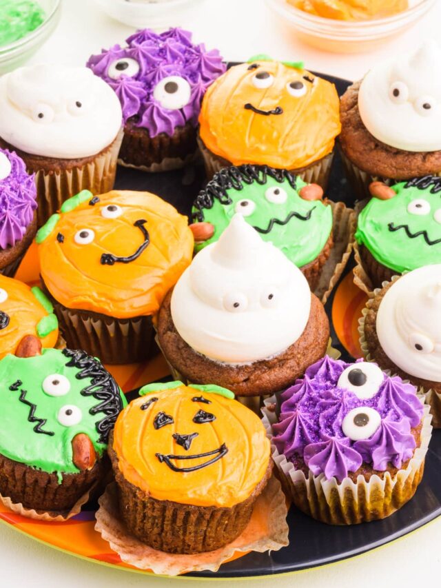 A plateful of colorful vegan halloween cupcakes sits in front of green and orange frosting bowls and googly eyes.