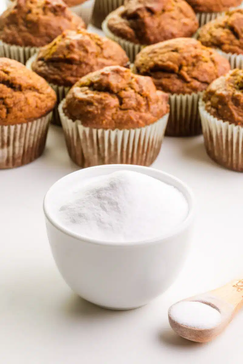 A bowl of baking soda sits in front of baked muffins in the background. There's a measuring spoon of baking soda beside the bowl.