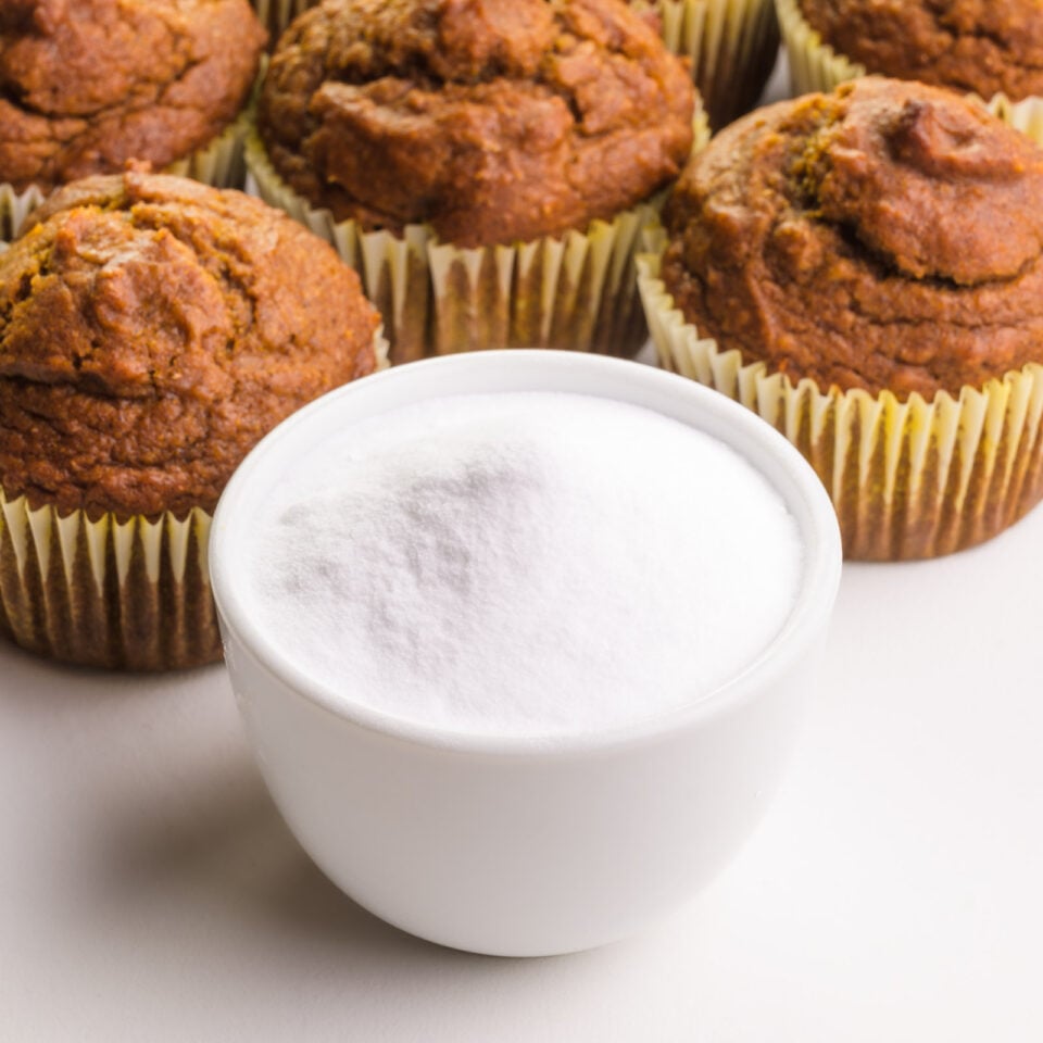 A spoonful of baking A bowl of baking soda sits in front of baked muffins in the background.