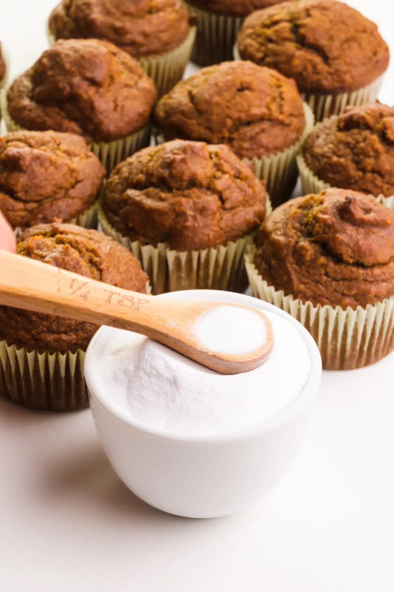 A spoonful of baking soda hovers over a bowl of it. There are baked muffins in the background.